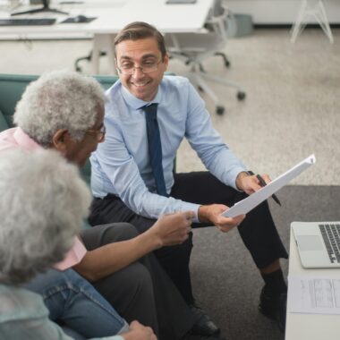 Consultant discussing financial plans with senior clients in a modern office setting, using documents and a laptop.